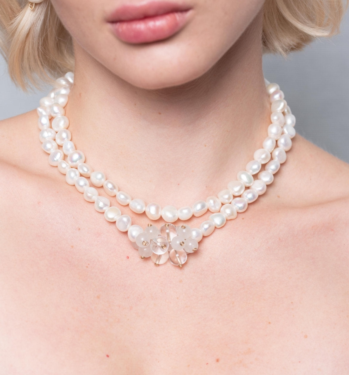 A close-up of a model wearing a double-strand pearl necklace with a central floral cluster of clear crystals and small white pearls.