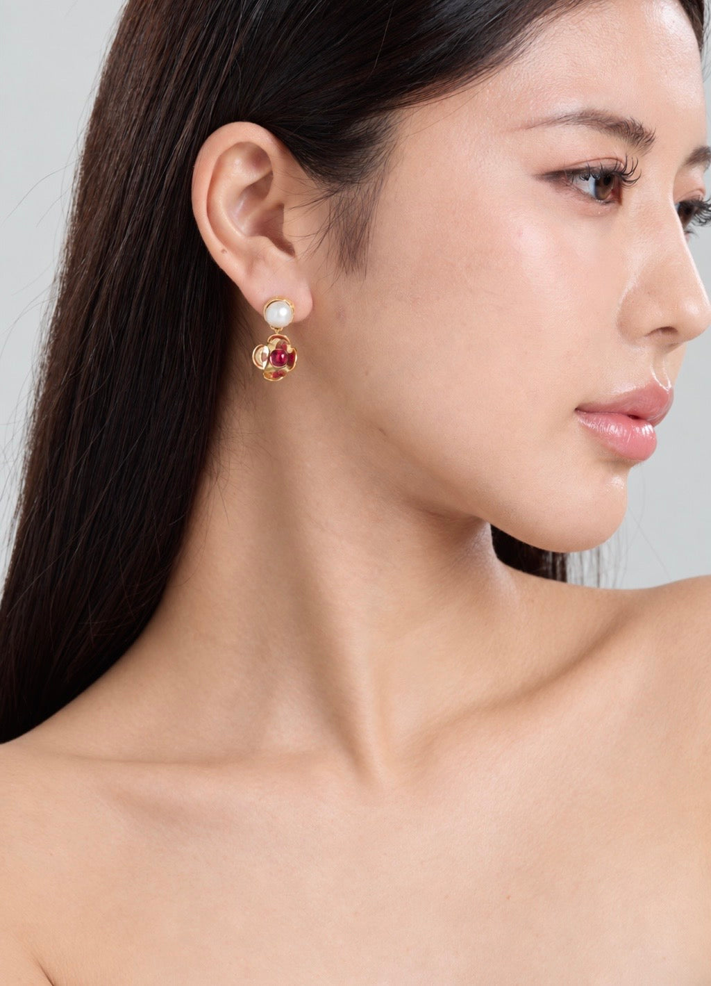 A close-up side profile of a woman wearing a gold flower shaped earring with a round red gemstone in the center, dangling from a white pearl stud top, set against a clean light gray background.
