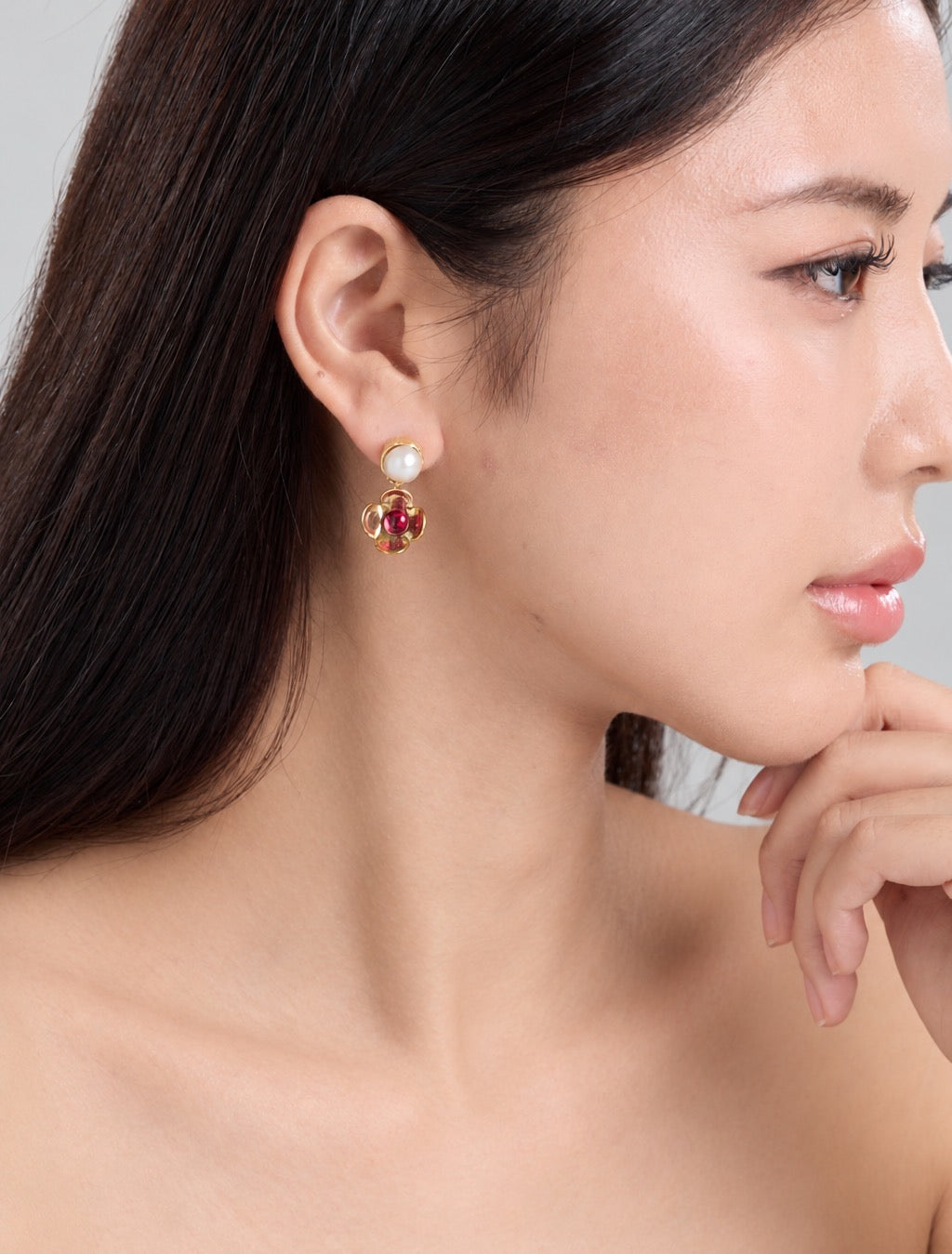 A close-up side profile of a woman wearing a gold and red floral drop earring with a white pearl stud above it, against a soft light gray background.