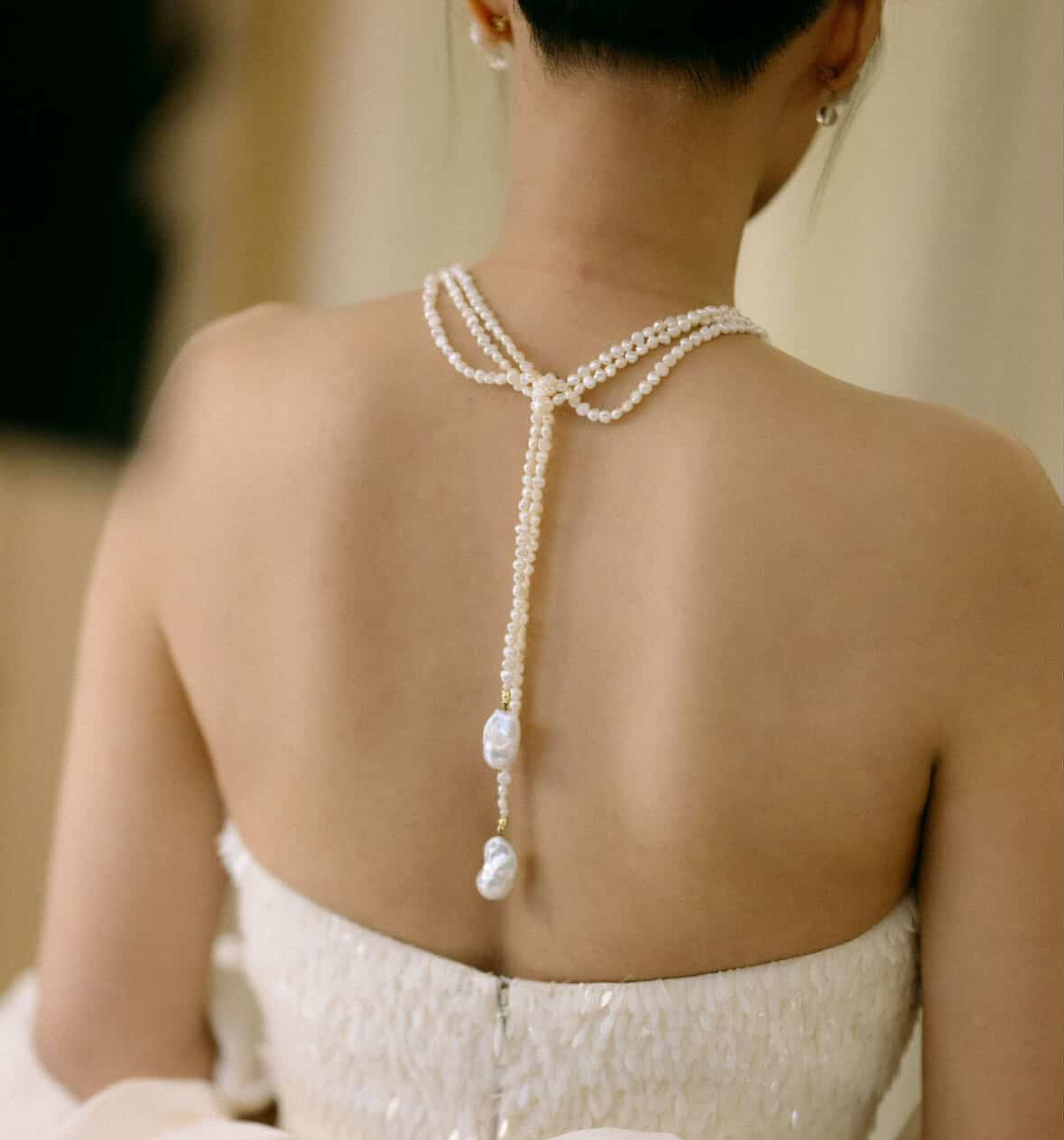 Close-up back view of a woman wearing a strapless white bridal gown with a pearl necklace with baroque pearls at each end, tied across her upper back.