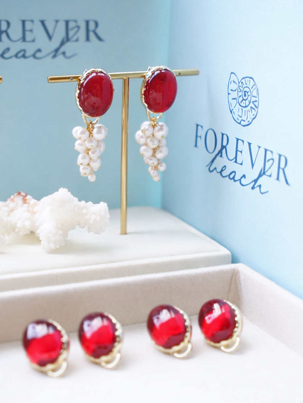 Red and pearl grape drop earrings displayed on a gold stand, with matching red and pearl stud earrings lined up in front on a jewelry box. The background features the Forever Beach logo on a light blue card.