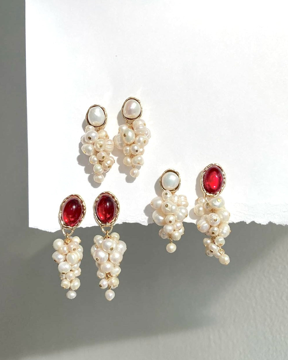 Three pairs of gold-tone dangle earrings with white pearl grape clusters and oval red gemstones, arranged on a white paper cutout against a gray wall.
