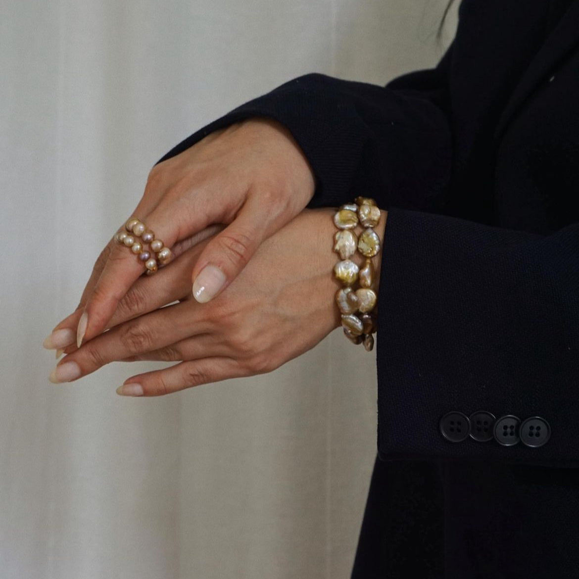 A close-up of a person's hands wearing a pearl baroque bracelet on one wrist and a matching multi-strand pearl ring on the other hand, styled with a dark navy blazer against a soft beige curtain background.