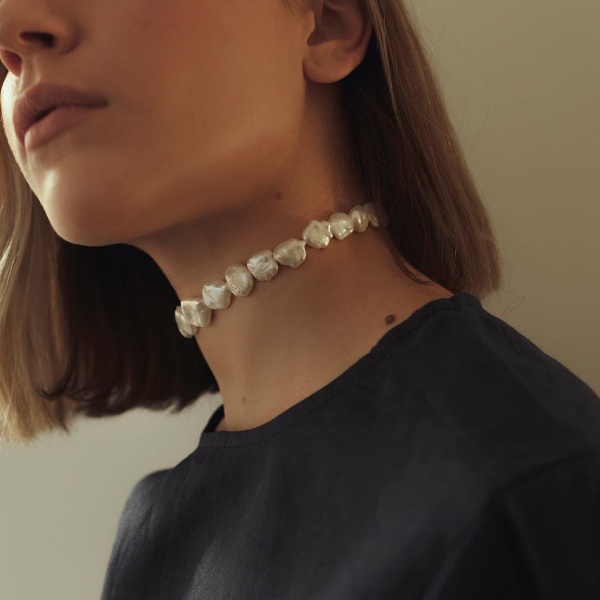 A close-up of a model wearing a delicate choker necklace made of irregular, organic-shaped white pearls, paired with a dark navy blue top.