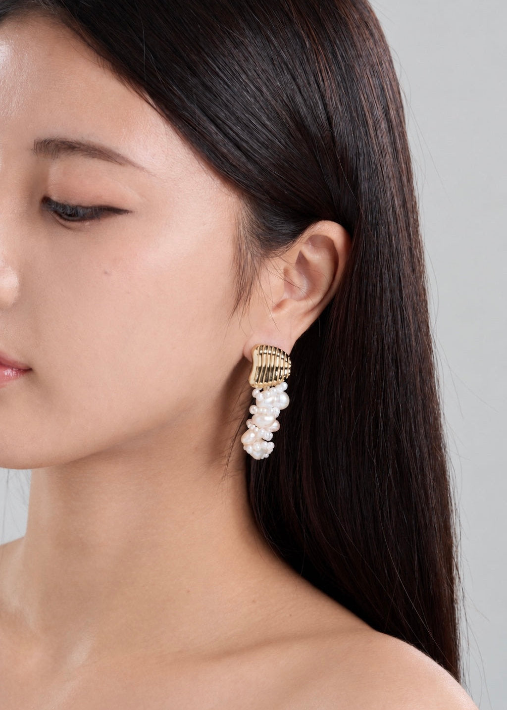 A close-up side profile of a woman with long dark hair wearing a gold and white drop earring with a textured gold top and a cluster of white pearl drops.