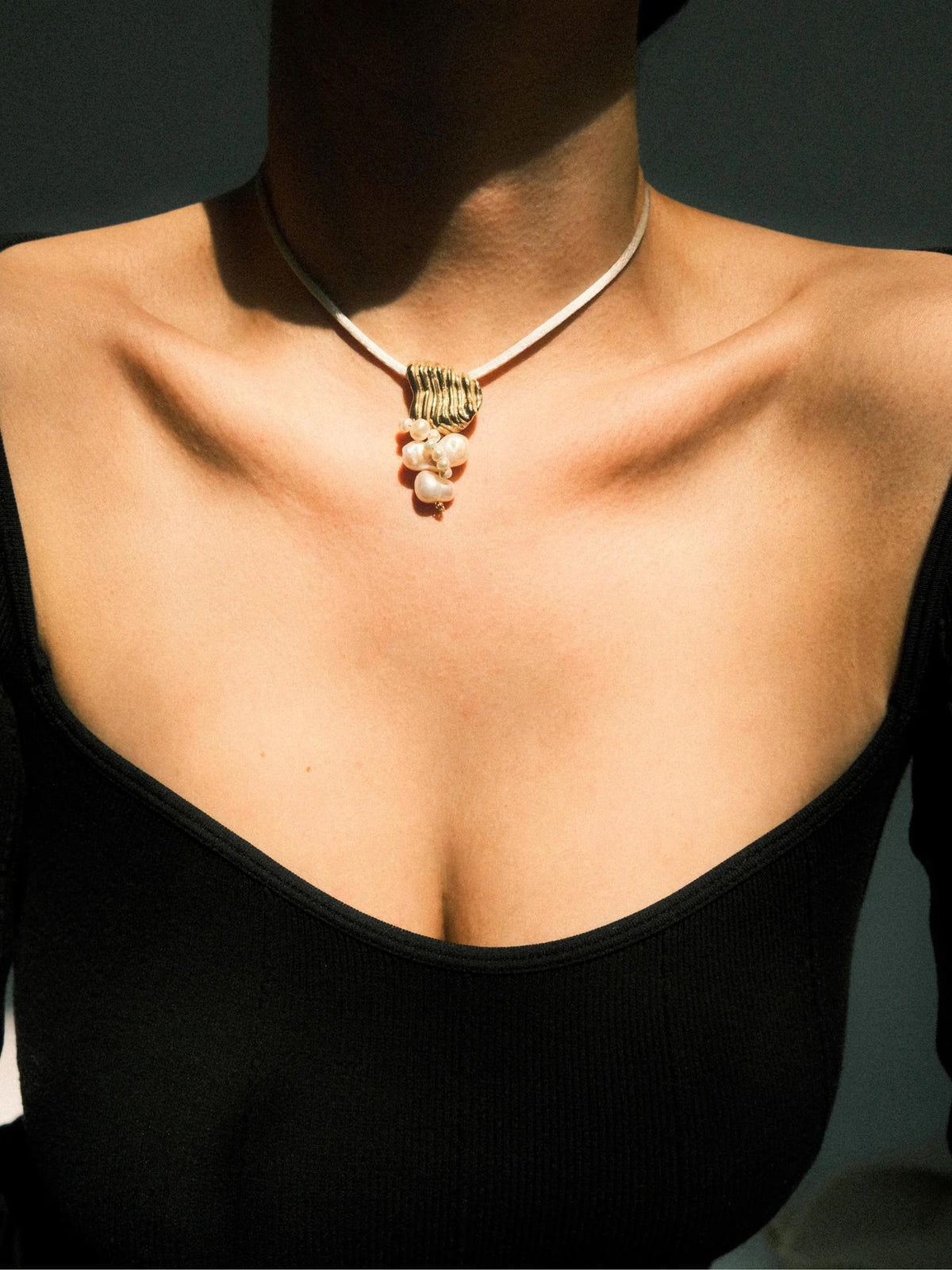A close-up of a model's neck and chest wearing a delicate necklace with a textured gold charm and a cluster of white pearls at the center, paired with a black ribbed top.