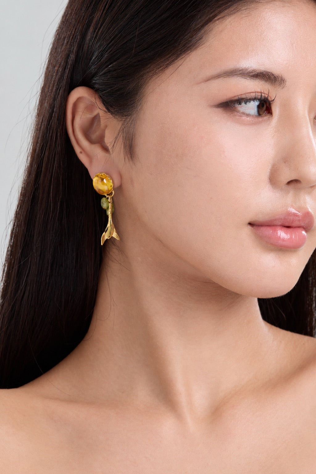 A close-up portrait of a woman with long dark hair wearing gold dangling earrings with amber-colored gemstone and leaf-like lower charms, against a light gray studio background.