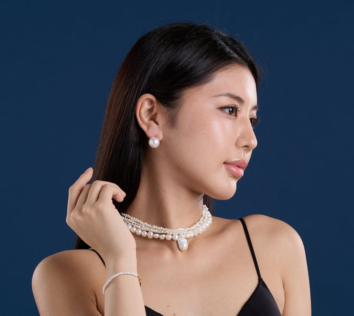 A woman in a black top models a multi-strand pearl necklace and matching pearl stud earring against a dark blue studio background.