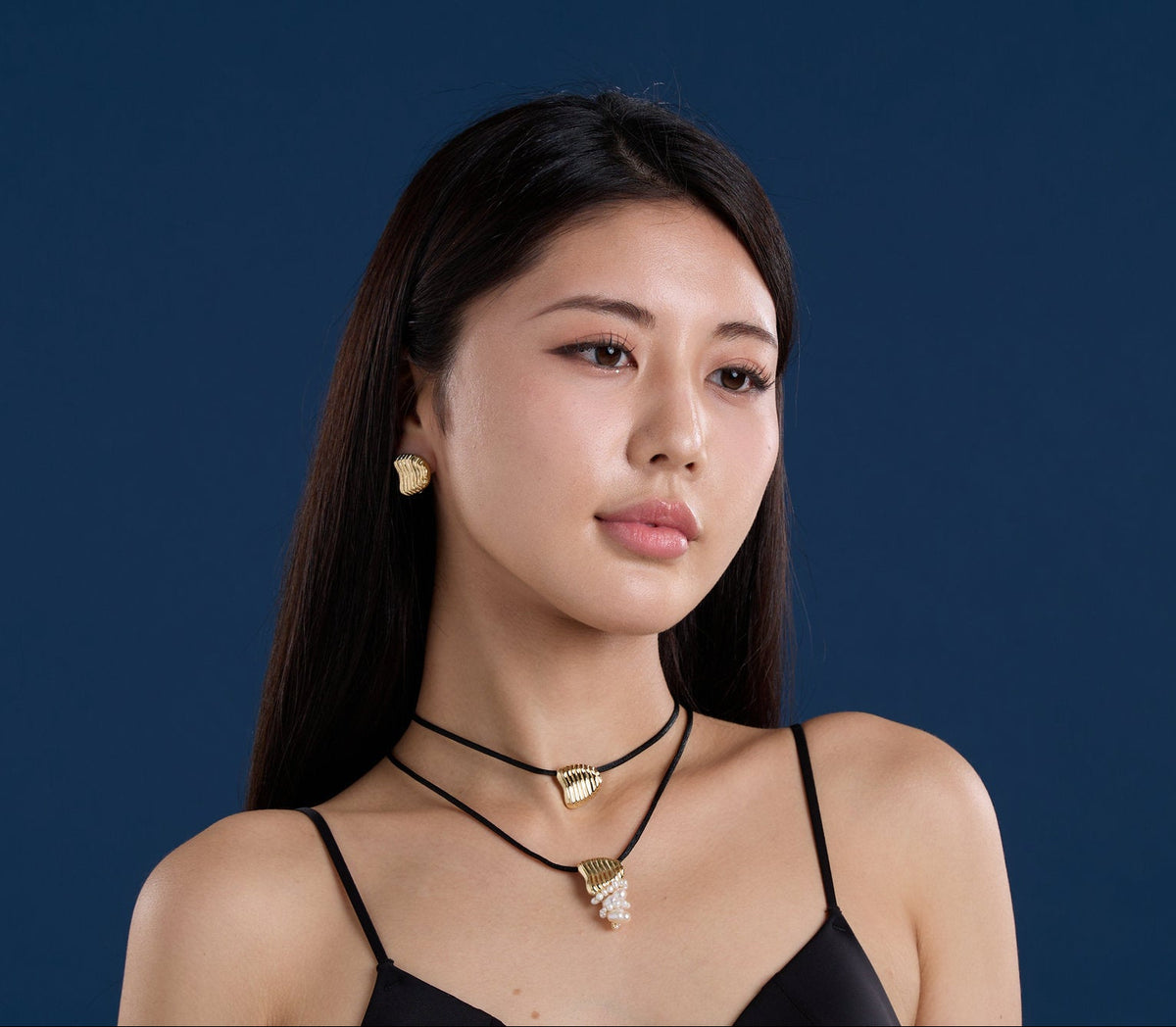 A woman with long dark hair is shown from the chest up against a solid dark blue background, wearing layered black cord necklaces with gold-toned pendants and matching gold stud earrings.